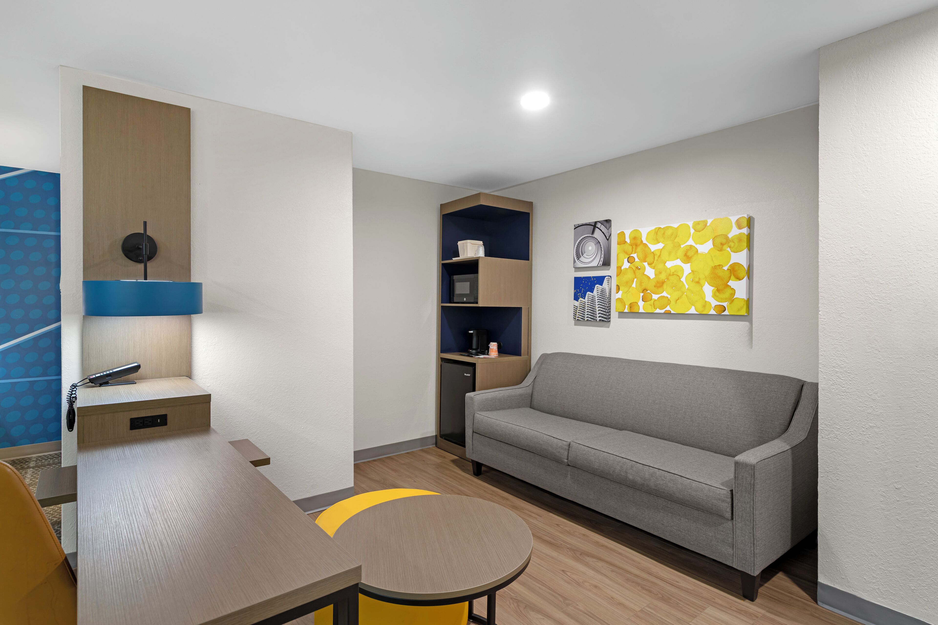 A modern hotel room with a sofa, a desk, a lamp, a coffee machine, and a wall art piece.