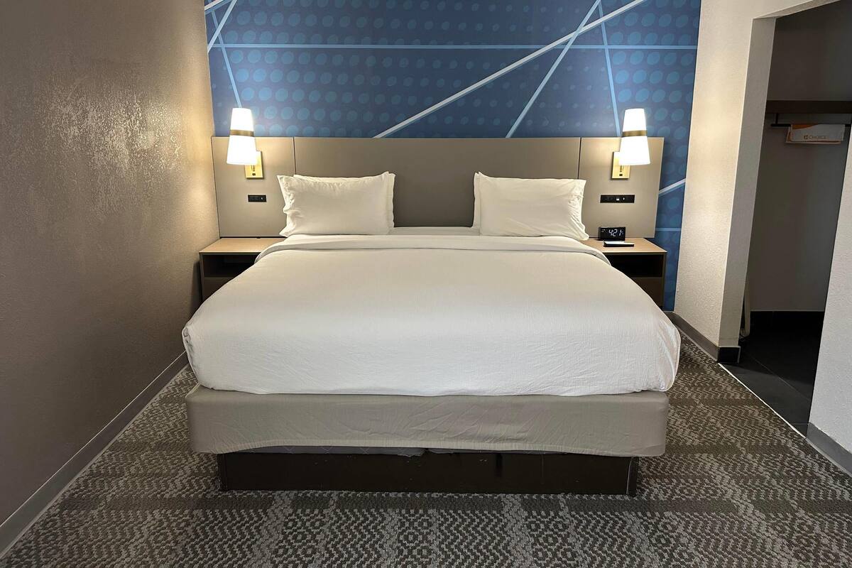 A hotel room with a large bed, two bedside lamps, a digital clock, and a patterned carpet.