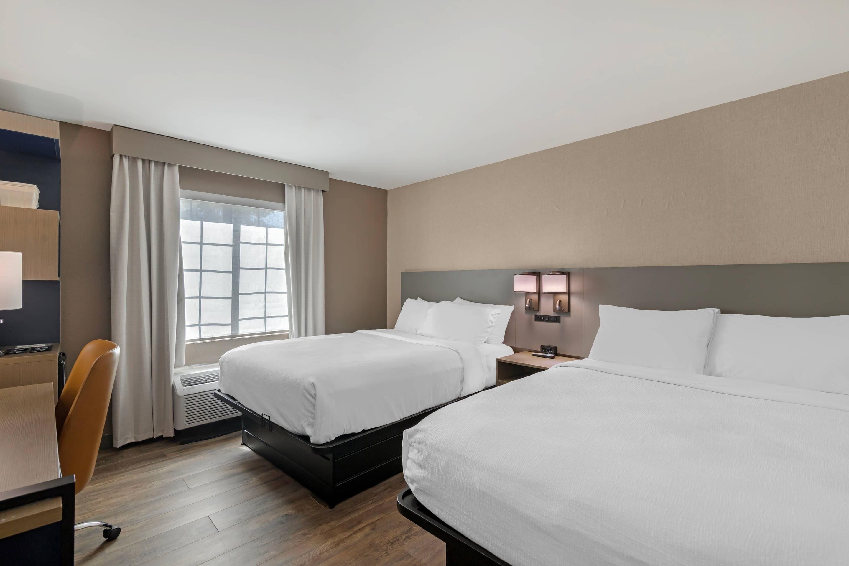 A hotel room with two beds, a desk, a chair, a window with curtains, and a wall-mounted lamp.