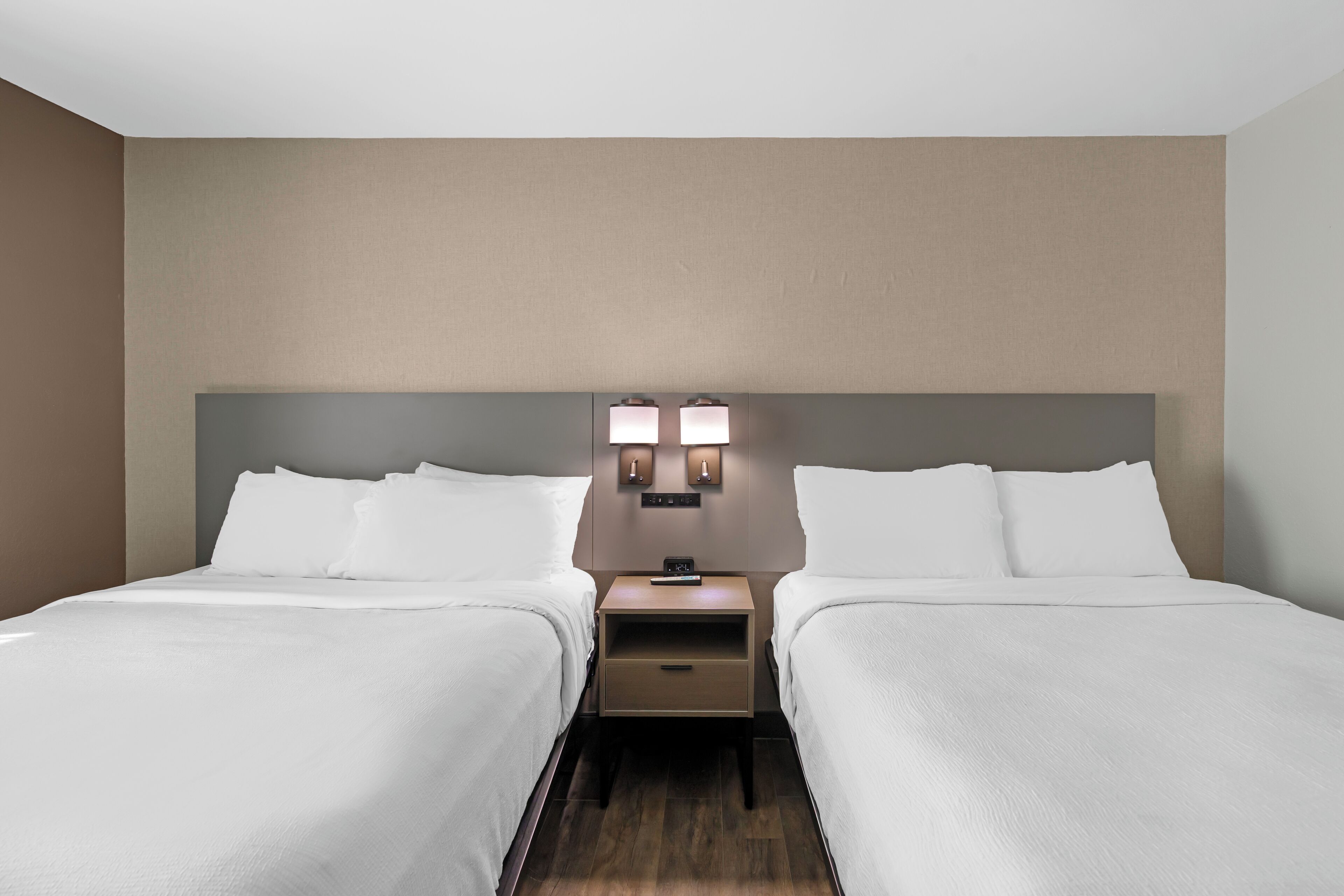 A hotel room with two beds, a nightstand with a lamp, and a neutral-colored wall.