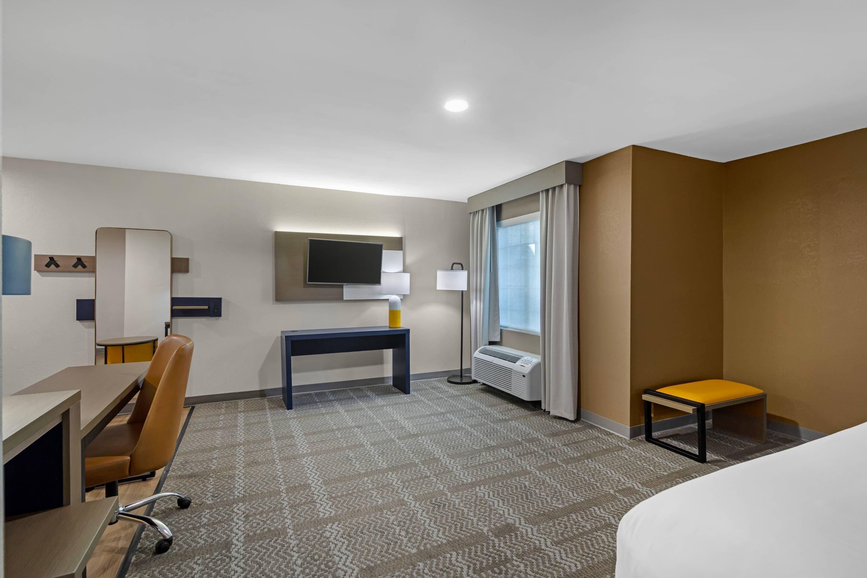 A hotel room with a bed, a desk, a chair, a TV, a lamp, a window with curtains, and a radiator.