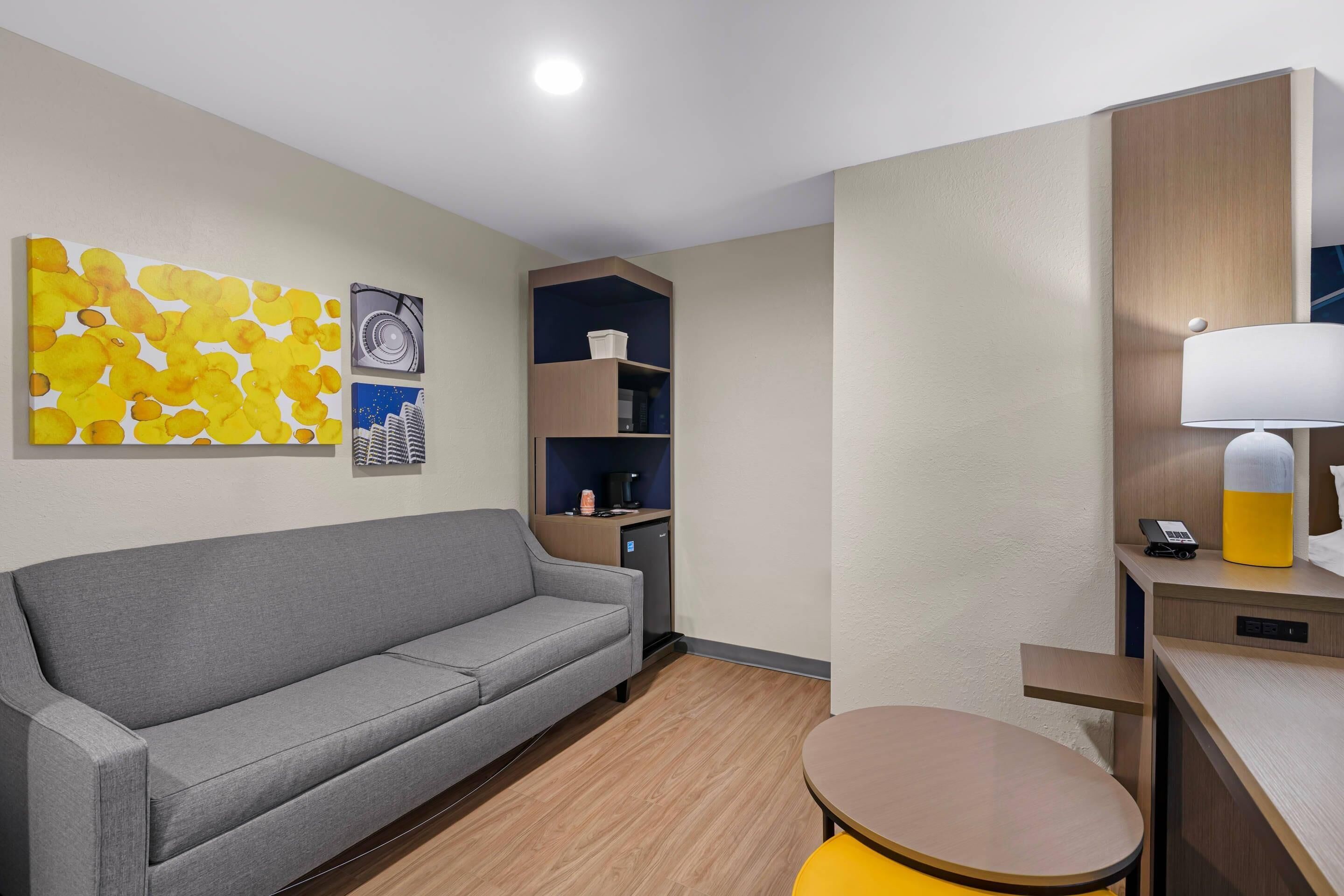 A hotel room with a grey sofa, a small round table, a lamp, a mini fridge, and a wall with abstract art and a cityscape.