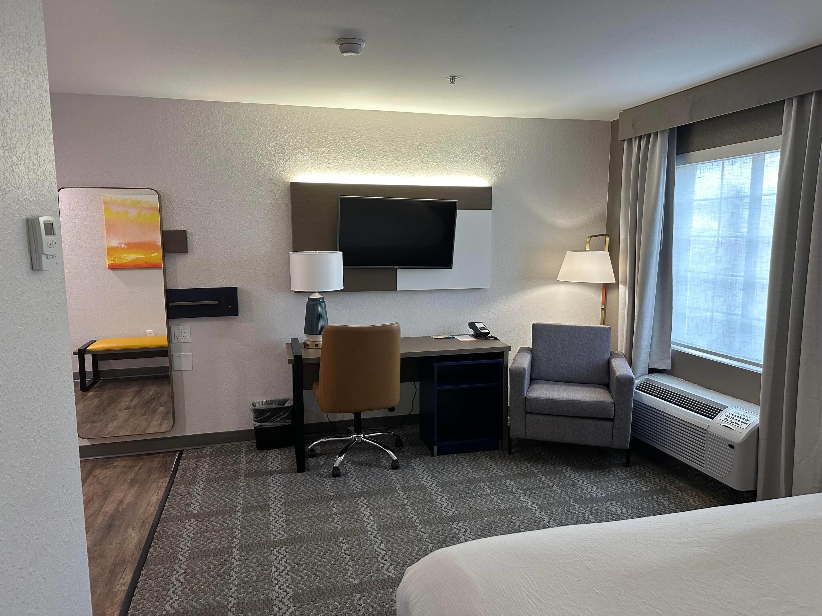 A hotel room with a bed, a desk with a chair, a TV, a lamp, and a window with curtains.
