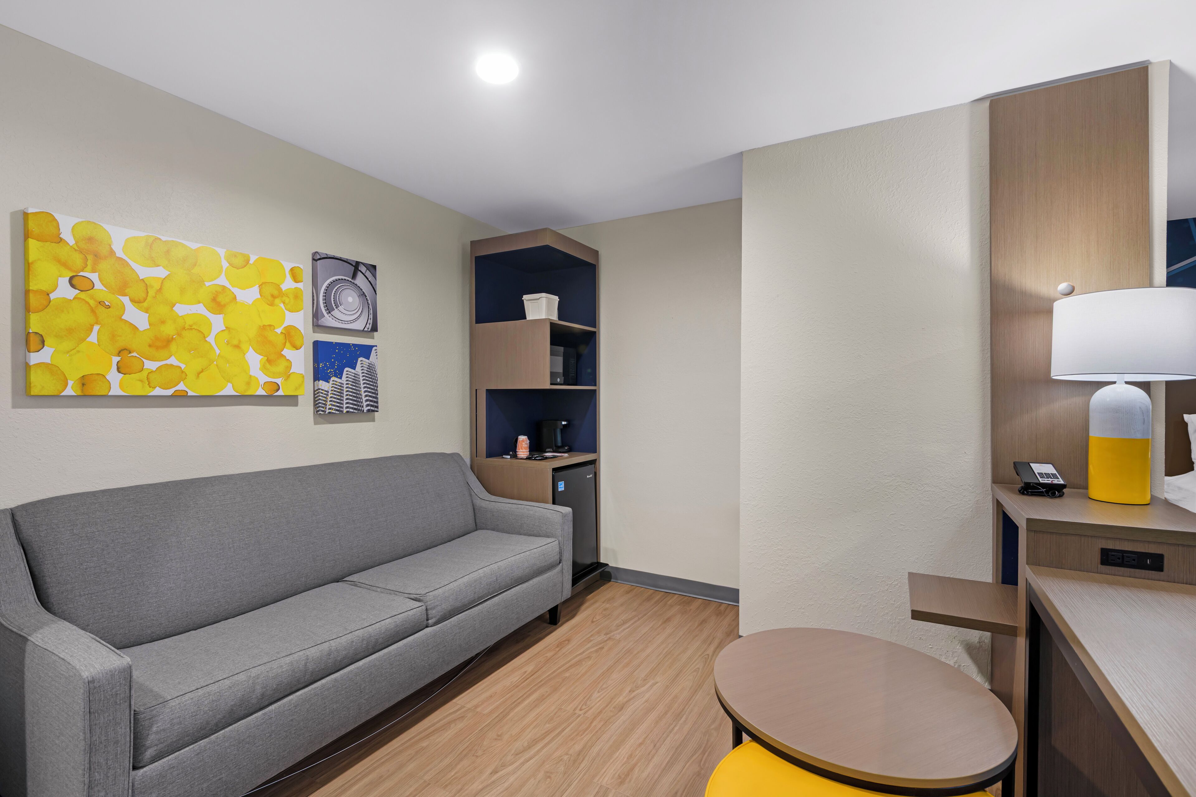 A modern hotel room with a grey sofa, a small round table, a lamp, a mini-fridge, and a built-in shelf unit.