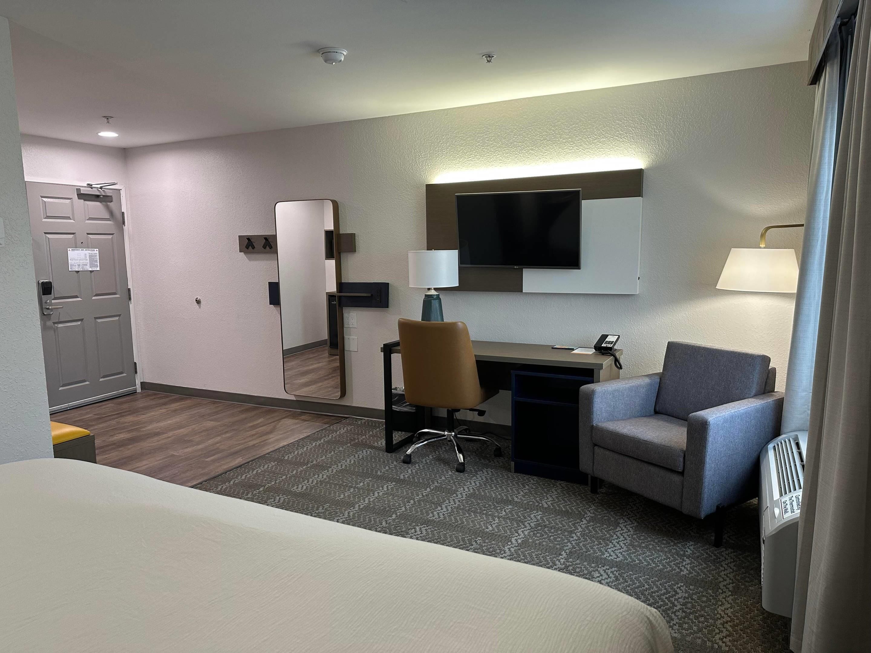 A hotel room with a bed, a desk, a chair, a television, a mirror, a lamp, and a radiator.