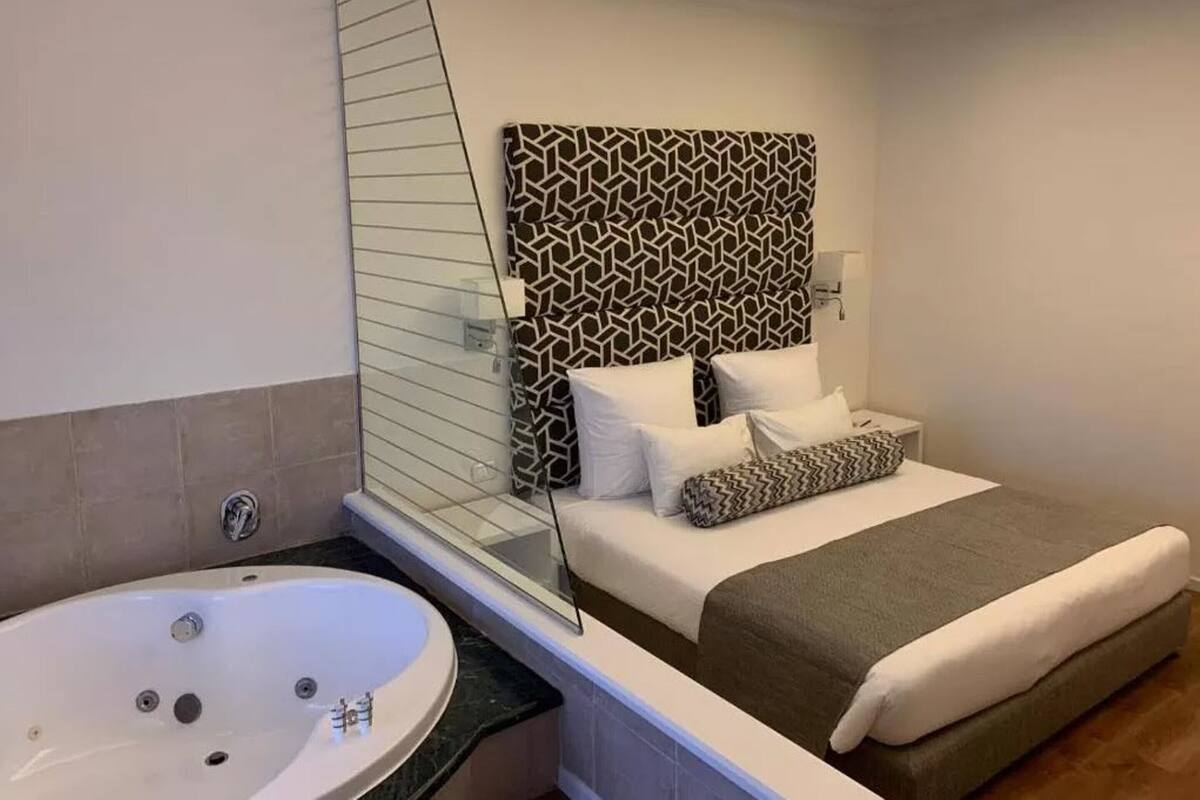Un baño moderno con una bañera grande, cabina de ducha de vidrio y una cama con cabecera y almohadones estampados.