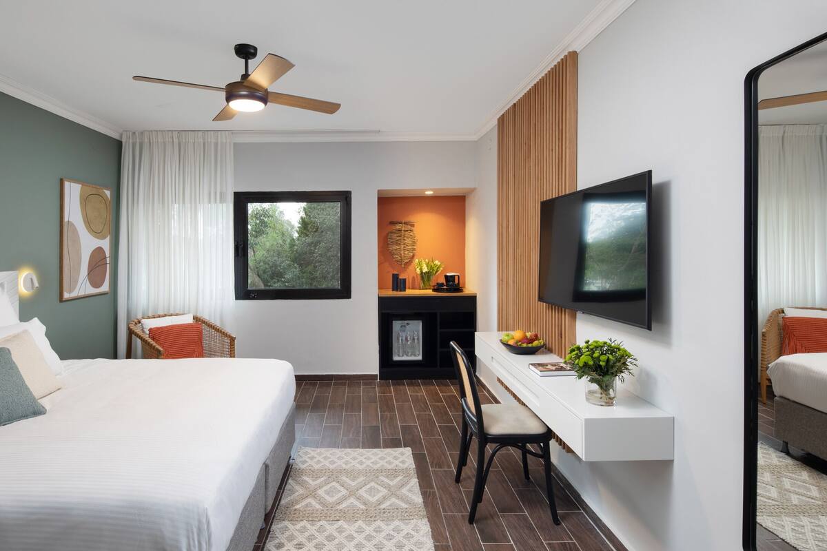 Una habitación de hotel moderna con una cama grande, un escritorio con televisor, un ventilador de techo y vistas a zonas verdes.