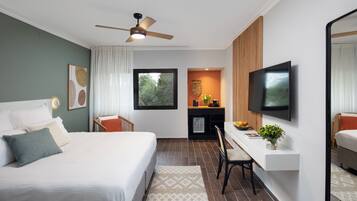 A modern hotel room with a large bed, a desk with a TV, a ceiling fan, and a view of greenery outside.