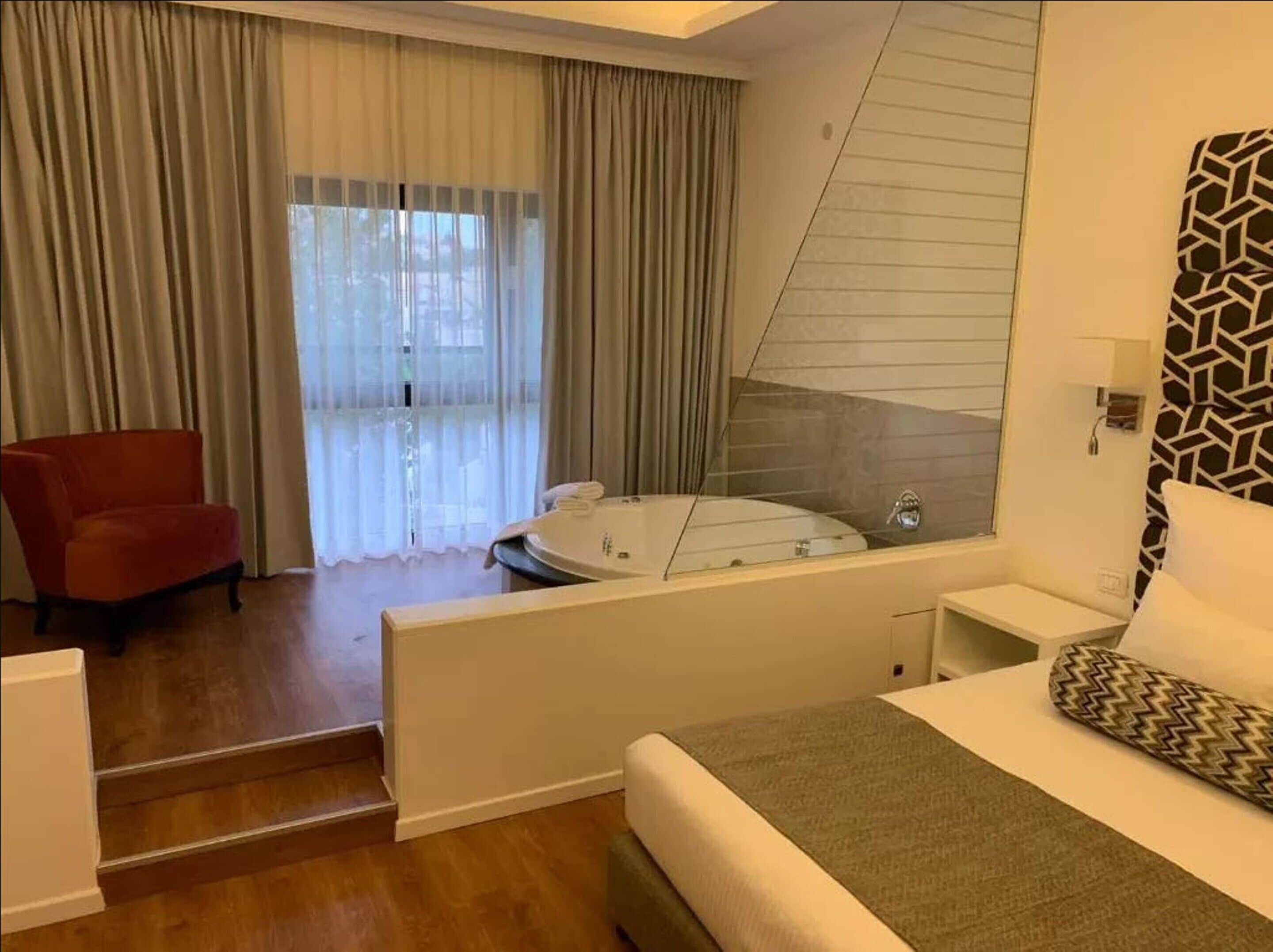 Una habitación de hotel con una cama, una silla roja, una bañera y un ventanal con cortinas.