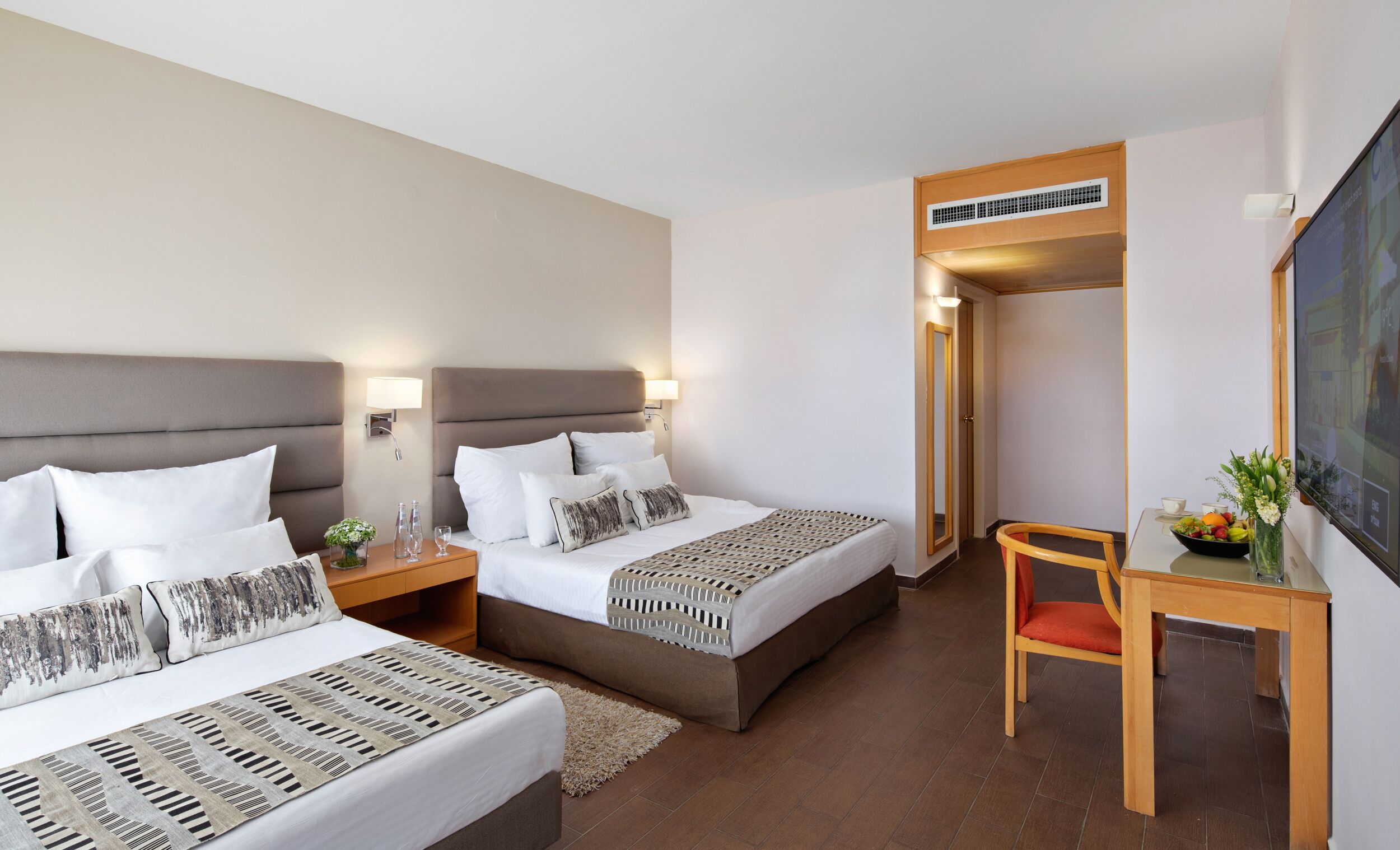 A hotel room with two beds, a desk, a chair, a TV, and a door leading to another room.