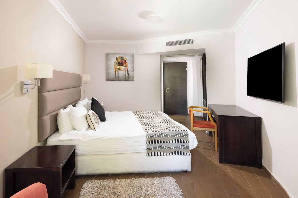 Una habitación de hotel moderna con cama, mesitas de noche, escritorio, silla, televisor de pantalla plana y una puerta que da a otra habitación.