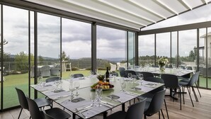 A dining area with tables set for a meal, large windows offering a view of the countryside, and a covered outdoor space.