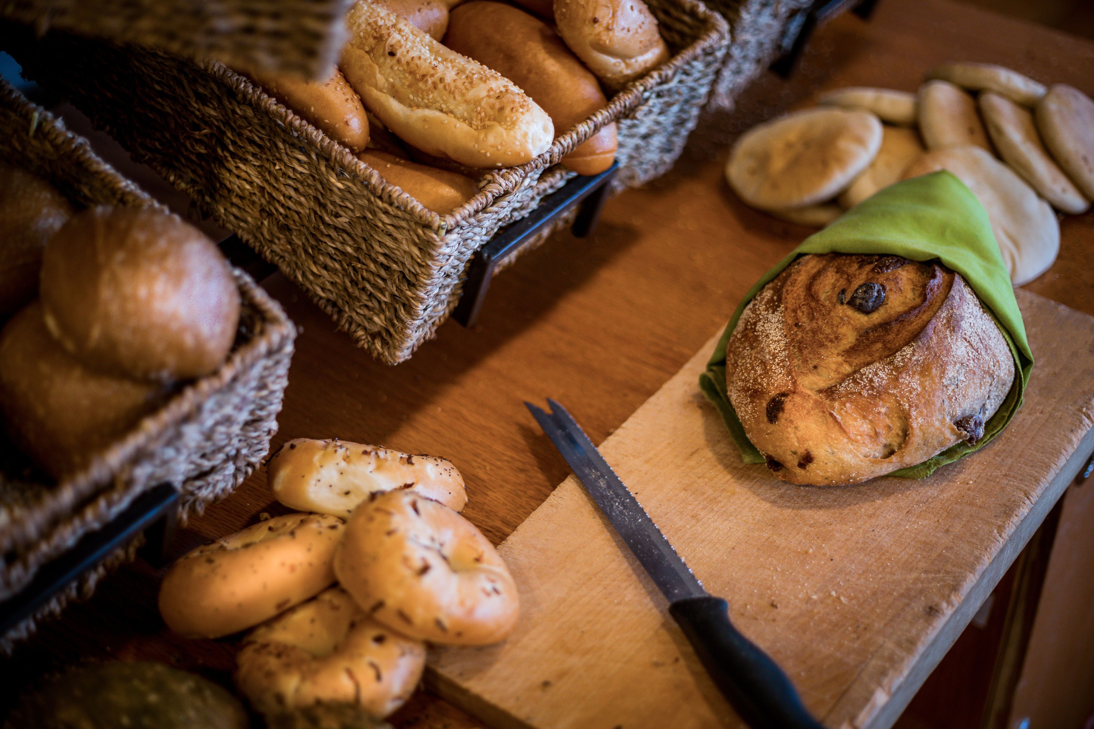 Verschiedene Brotsorten und Gebäck auf einem Holztisch, darunter ein Korb mit Laugen und ein Schneidebrett mit Messer.