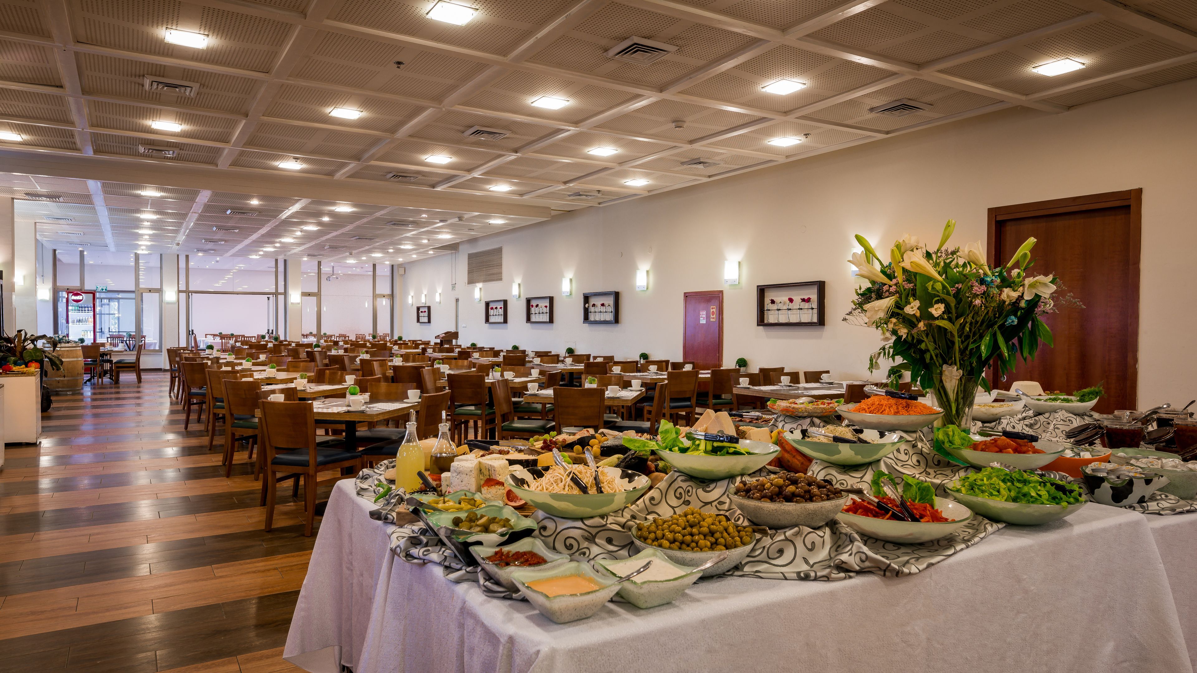 A spacious dining hall with multiple tables set for a buffet, featuring a variety of food items and a large floral centerpiece.