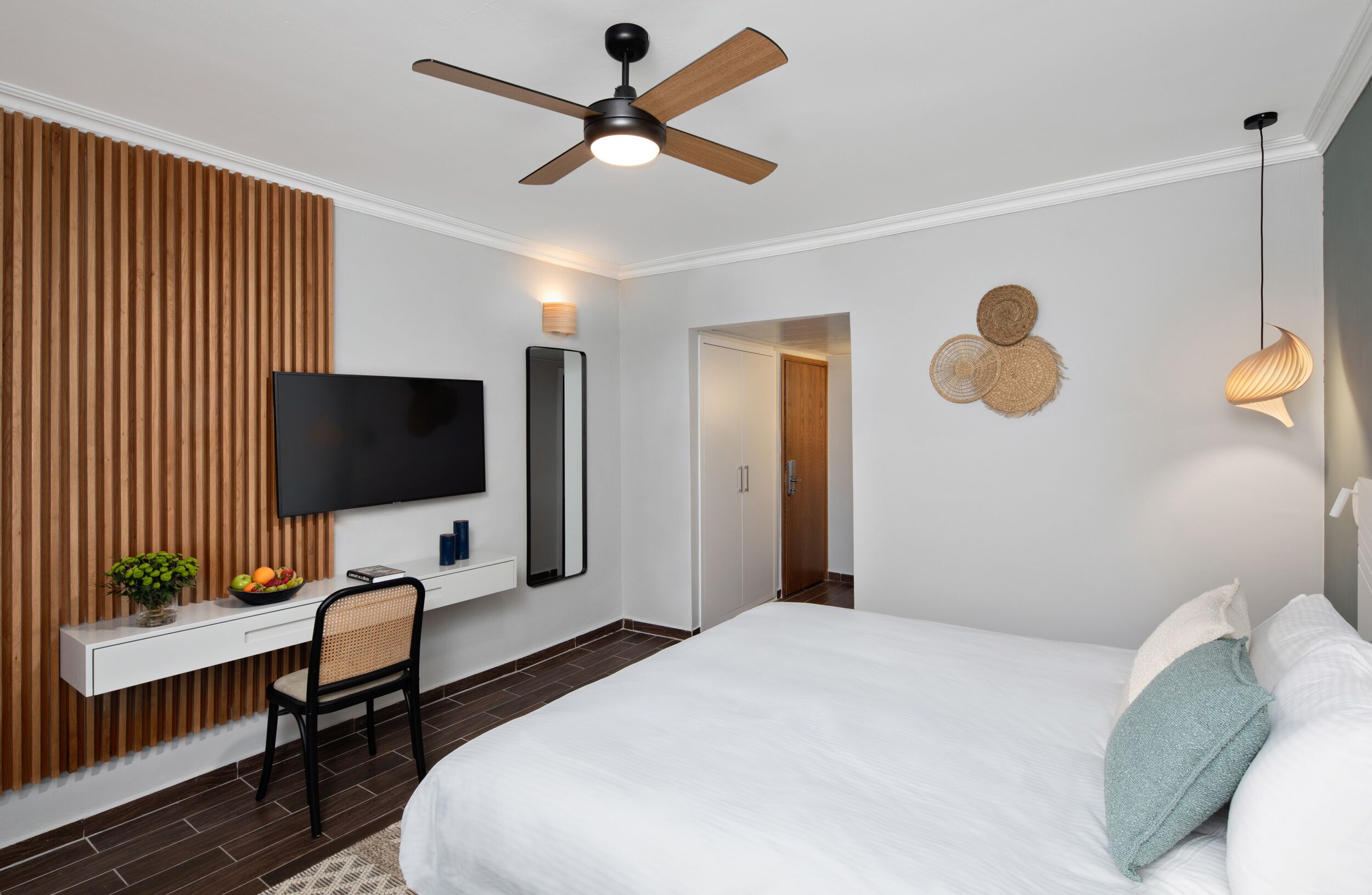 Et moderne hotelværelse med seng, skrivebord med stol, fjernsyn, loftsvifte og vægmonteret lampe.