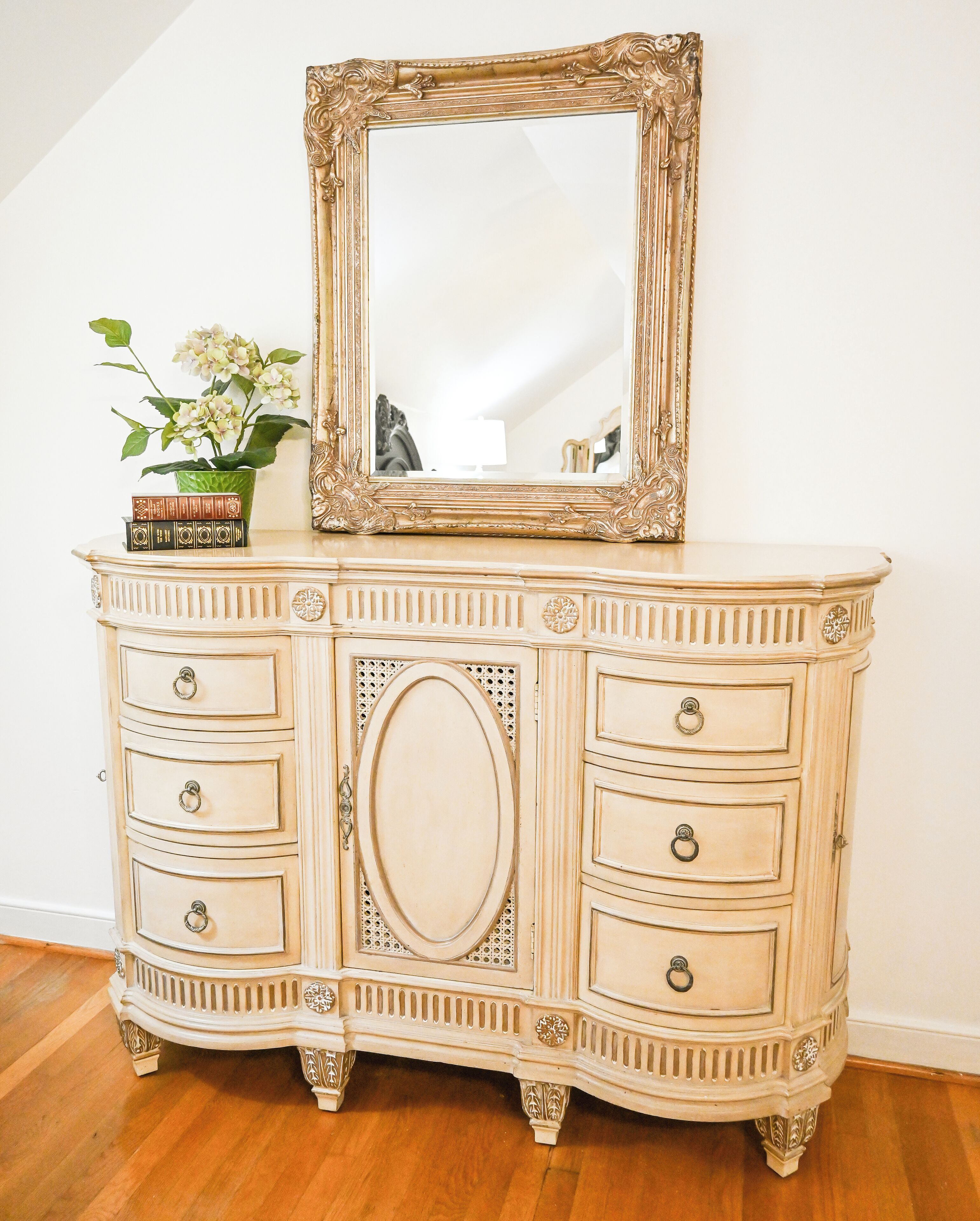 Une commode de couleur crème, richement décorée, dotée de nombreux tiroirs et surmontée d’un grand miroir.