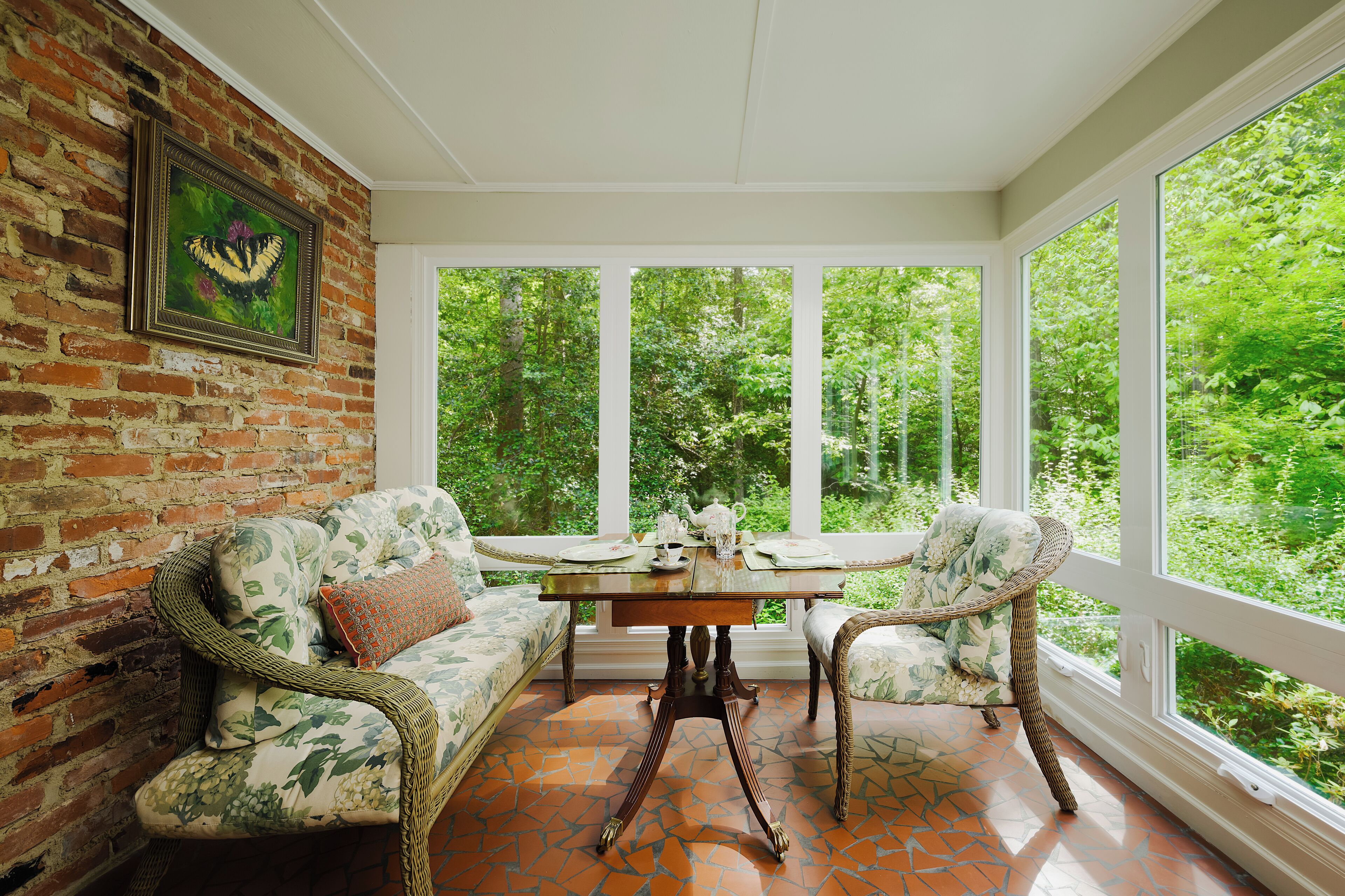 A sunroom with a brick wall, a framed butterfly artwork, a wooden table, and wicker furniture.