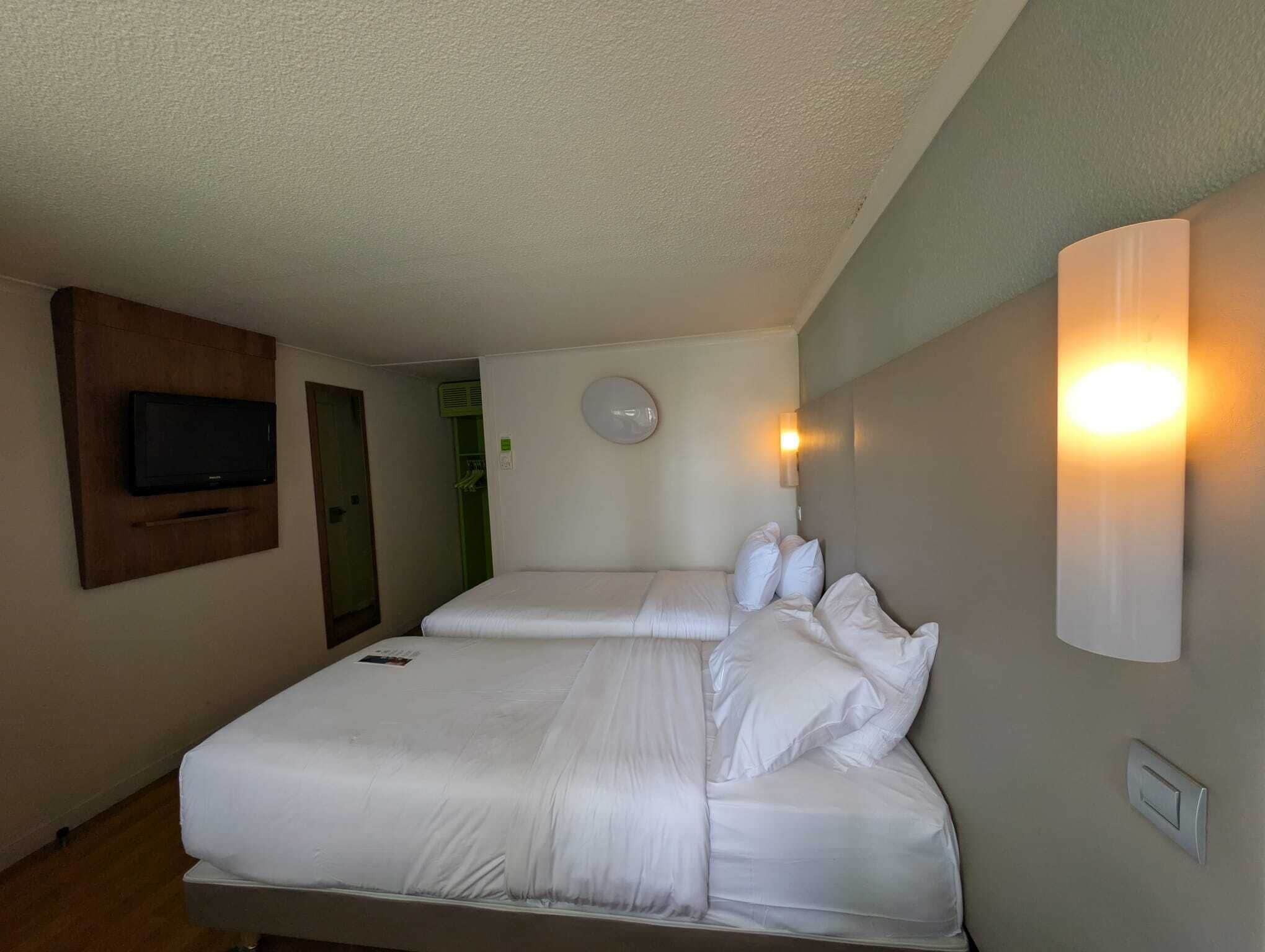 Habitación de hotel con dos camas, un televisor fijado a la pared, una lámpara en la pared y una puerta que da a un baño.