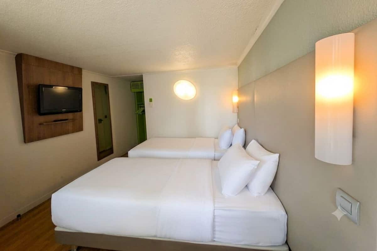 Quarto de hotel com duas camas, televisão, luminárias fixadas na parede e uma porta.