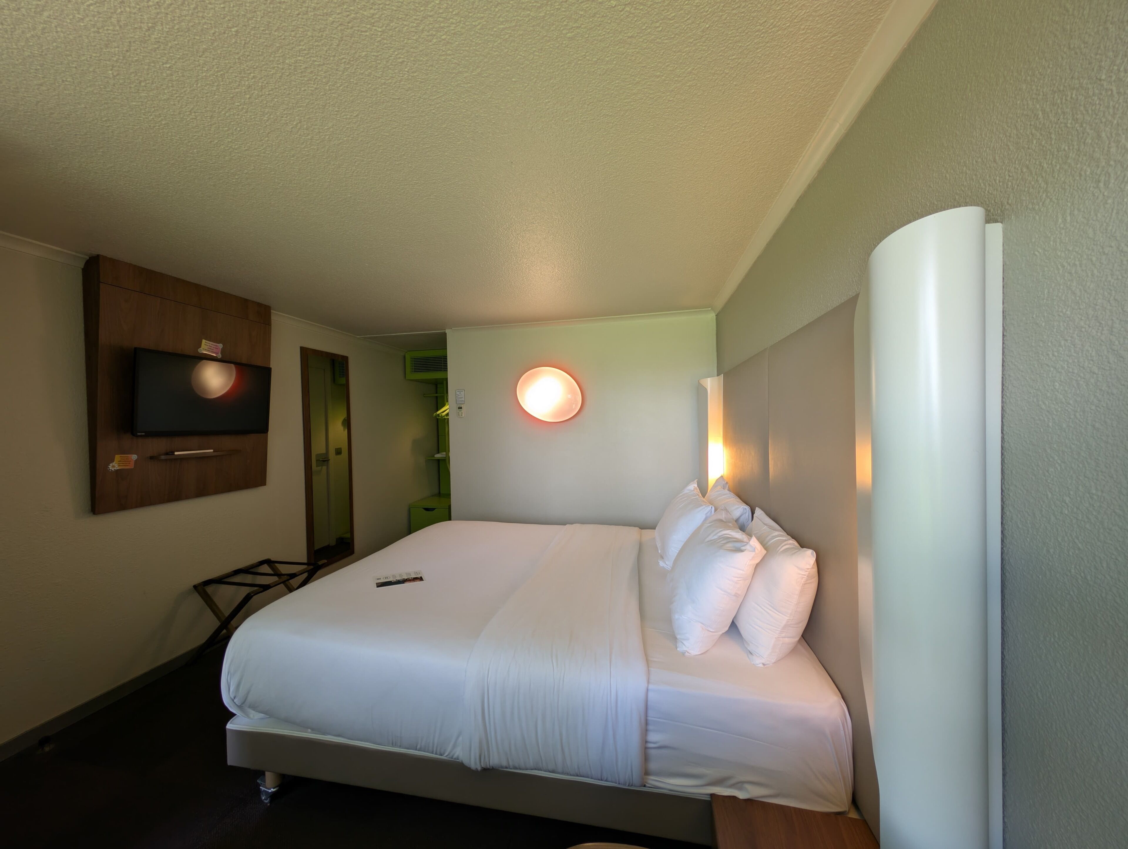 Una habitación de hotel con una cama, un televisor de pantalla plana montado en la pared, una mesita de noche y un armario verde.