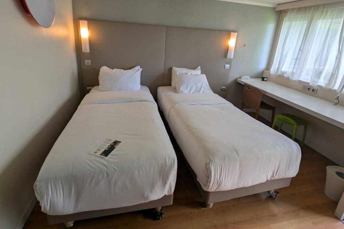 Ein Hotelzimmer mit zwei Einzelbetten, einem Schreibtisch, einem Stuhl und einem Fenster mit Vorhängen.