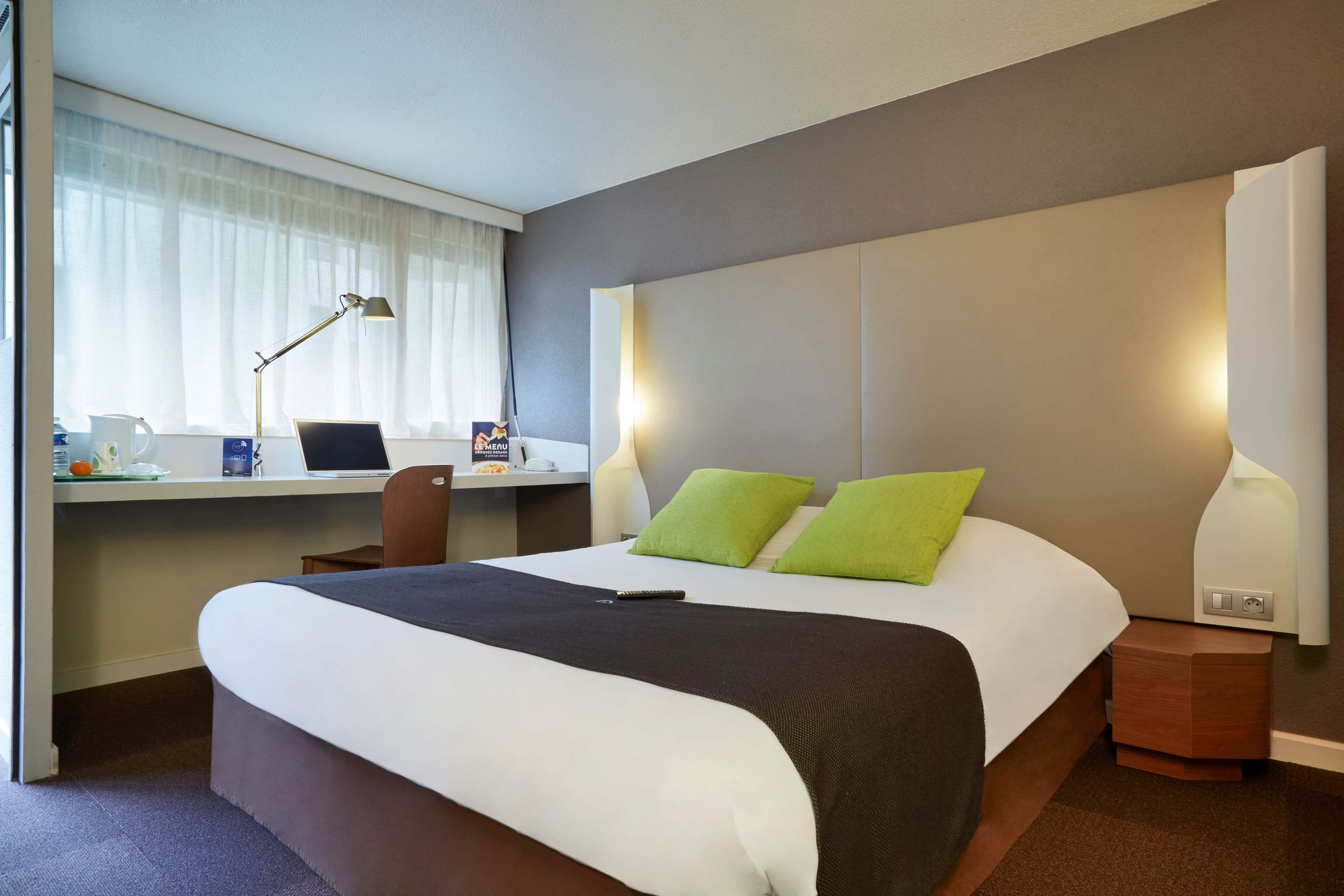 A hotel room with a bed, a desk with a laptop, a chair, and a bedside table with a lamp.