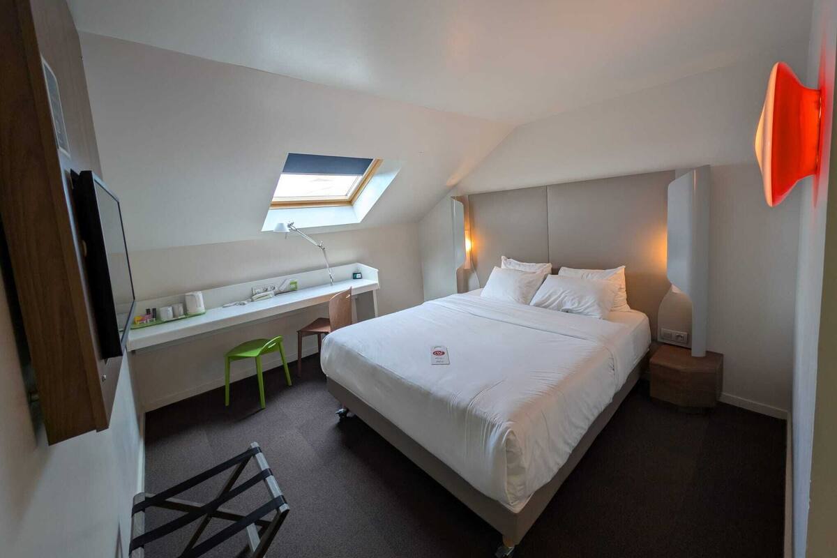 Ein Schlafzimmer mit einem Bett, einem Schreibtisch, einem Stuhl, einem Oberlicht und einer roten Wand als Blickfang.