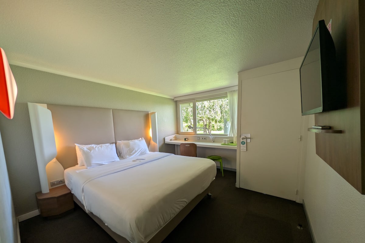 Quarto de hotel com cama, mesa de cabeceira, luminária, escrivaninha, cadeira, janela e televisão fixada na parede.