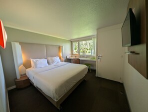 Quarto de hotel com cama, mesa de cabeceira, luminária, escrivaninha, cadeira, janela e televisão fixada na parede.