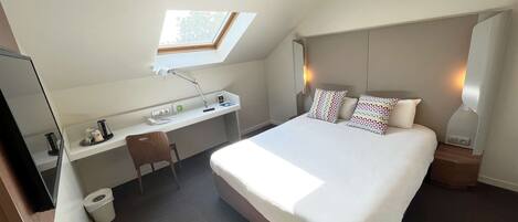Quarto de hotel com cama, escrivaninha, cadeira e claraboia.