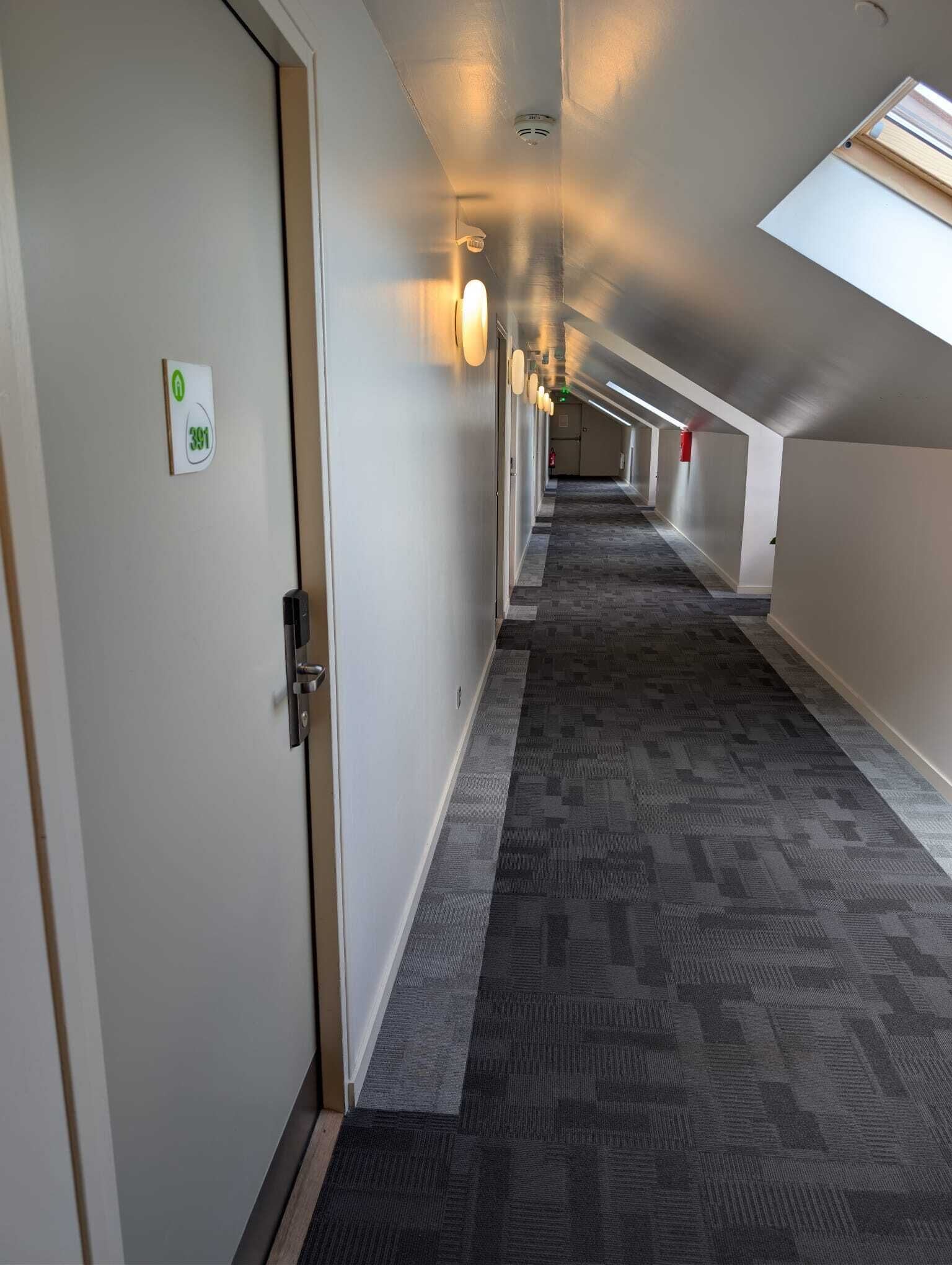 Un pasillo de hotel con piso alfombrado, una puerta con letrero verde y un tragaluz.