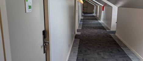 Un pasillo de hotel con piso alfombrado, una puerta con letrero verde y un tragaluz.