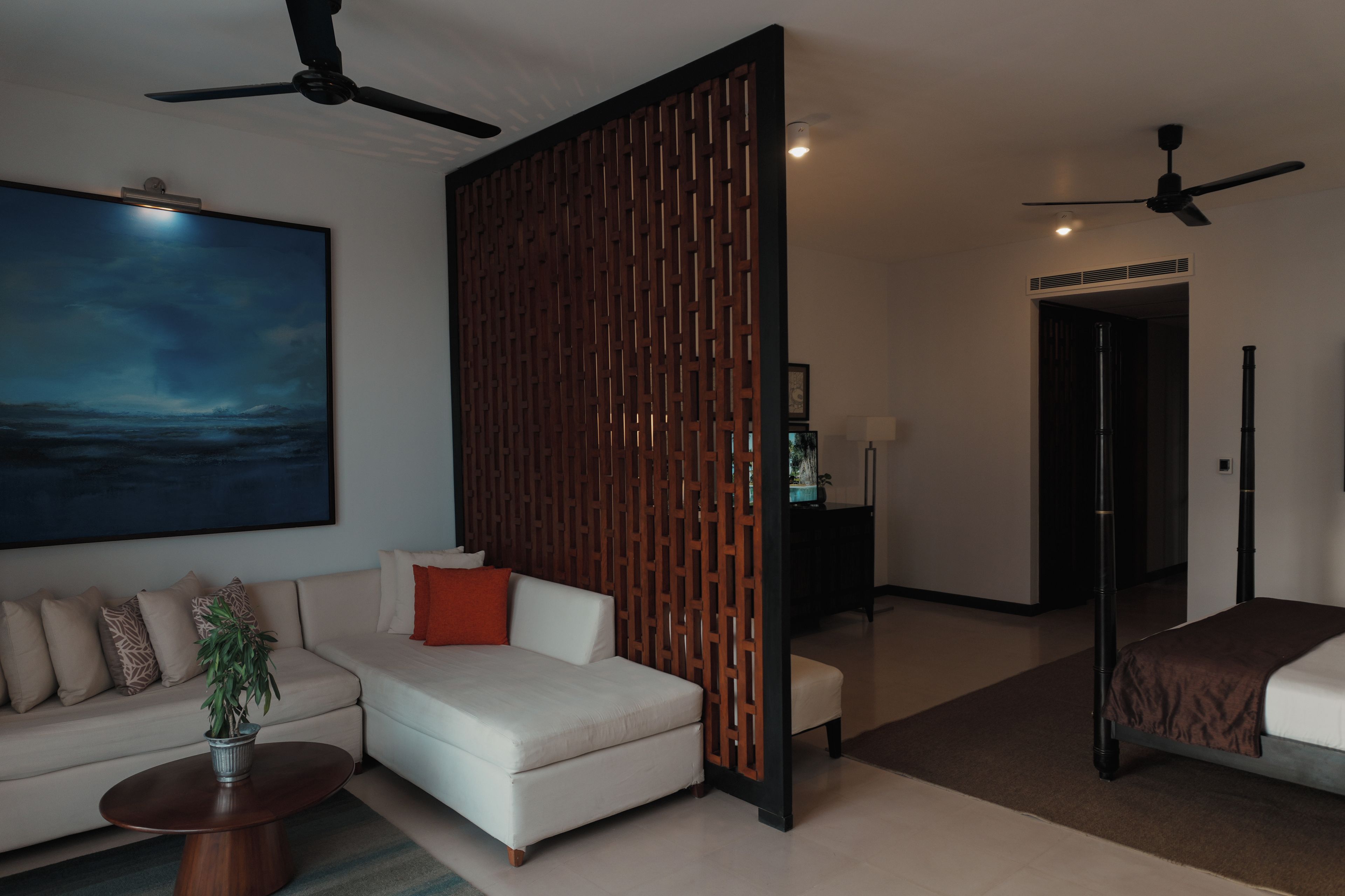 Una habitación de hotel moderna con un sofá modular, una mesa redonda de centro, un cuadro grande, un panel decorativo de madera y ventiladores de techo.
