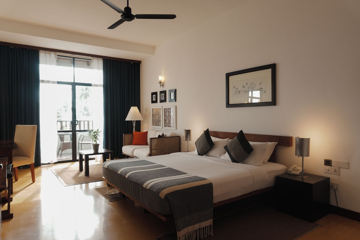 A hotel room with a bed, bedside tables, a desk, a chair, a lamp, a ceiling fan, and a window with curtains.