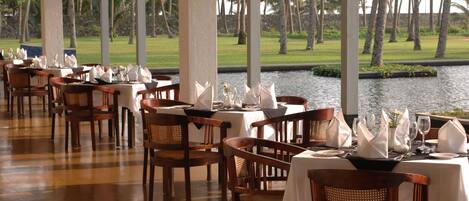 A restaurant with wooden chairs and tables, a view of palm trees, and a body of water in the background.
