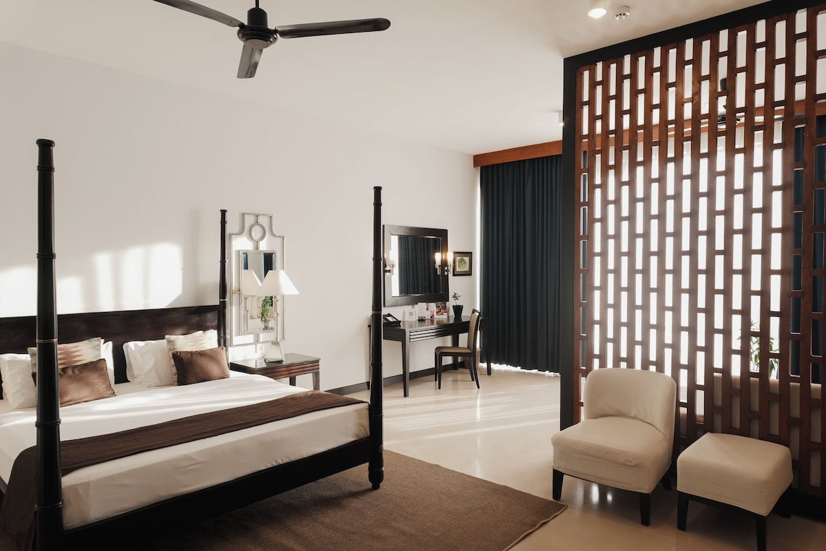 A four-poster bed with a dark headboard, a desk with a chair, a large mirror, and a partition with a lattice design.