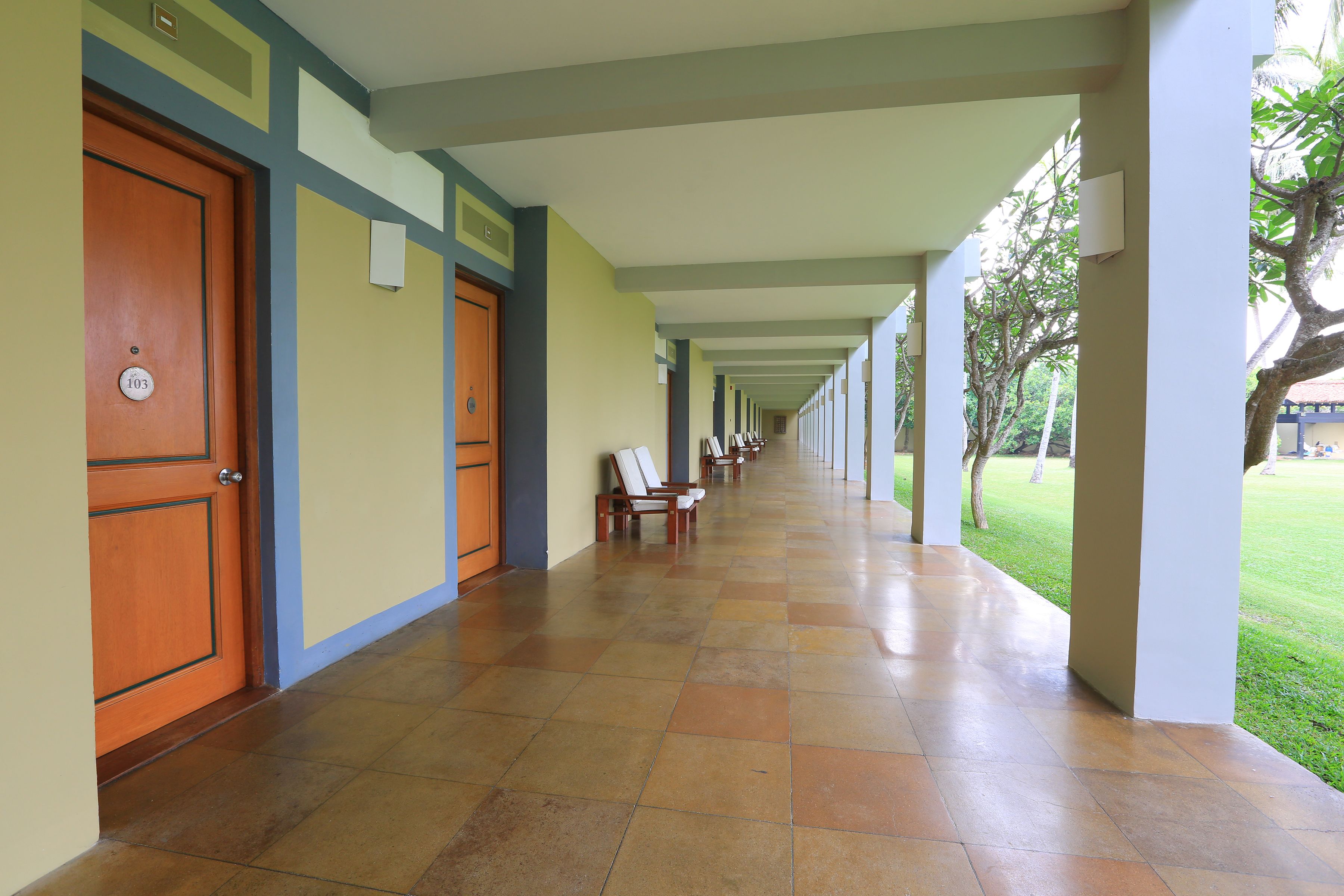 A long corridor with doors on the left, chairs along the right, and a covered area with a view of a grassy area.