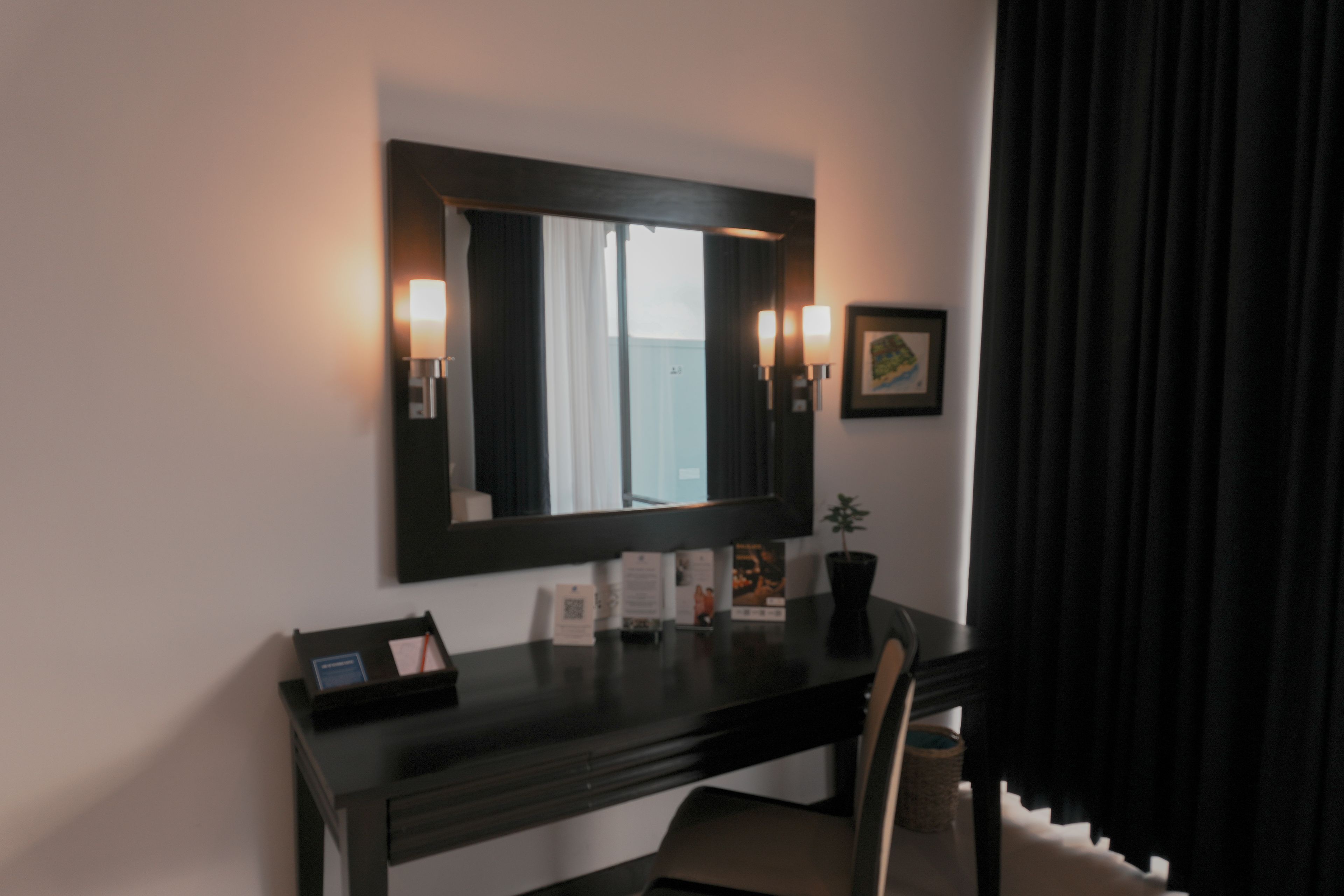 A hotel room with a dark wooden desk, a mirror, two wall-mounted lamps, a framed picture, and a small potted plant.
