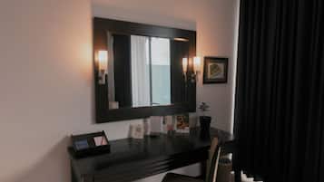 A hotel room with a dark wooden desk, a mirror, two wall-mounted lamps, a framed picture, and a small potted plant.
