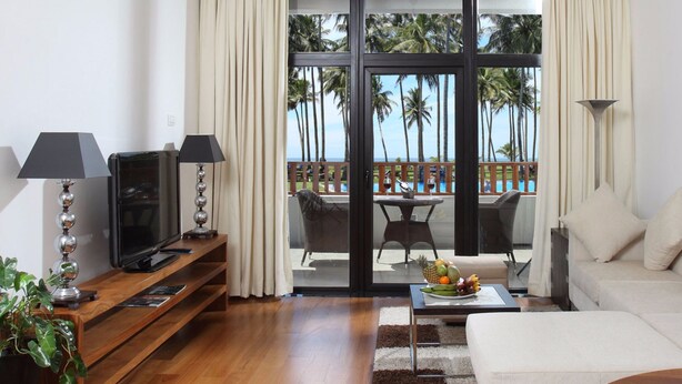 A modern living room with a TV, sofa, and dining area with a view of palm trees. - The Blue Water - Geoffrey Bawa Heritage Resort Hotel (Wadduwa)