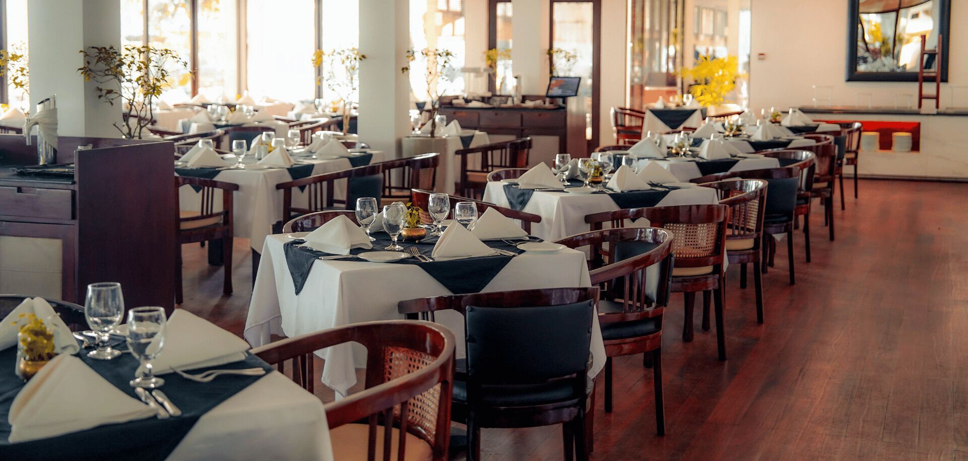 Een restaurant met meerdere tafels gedekt voor diner, elk met witte tafelkleden en donkere servetten.