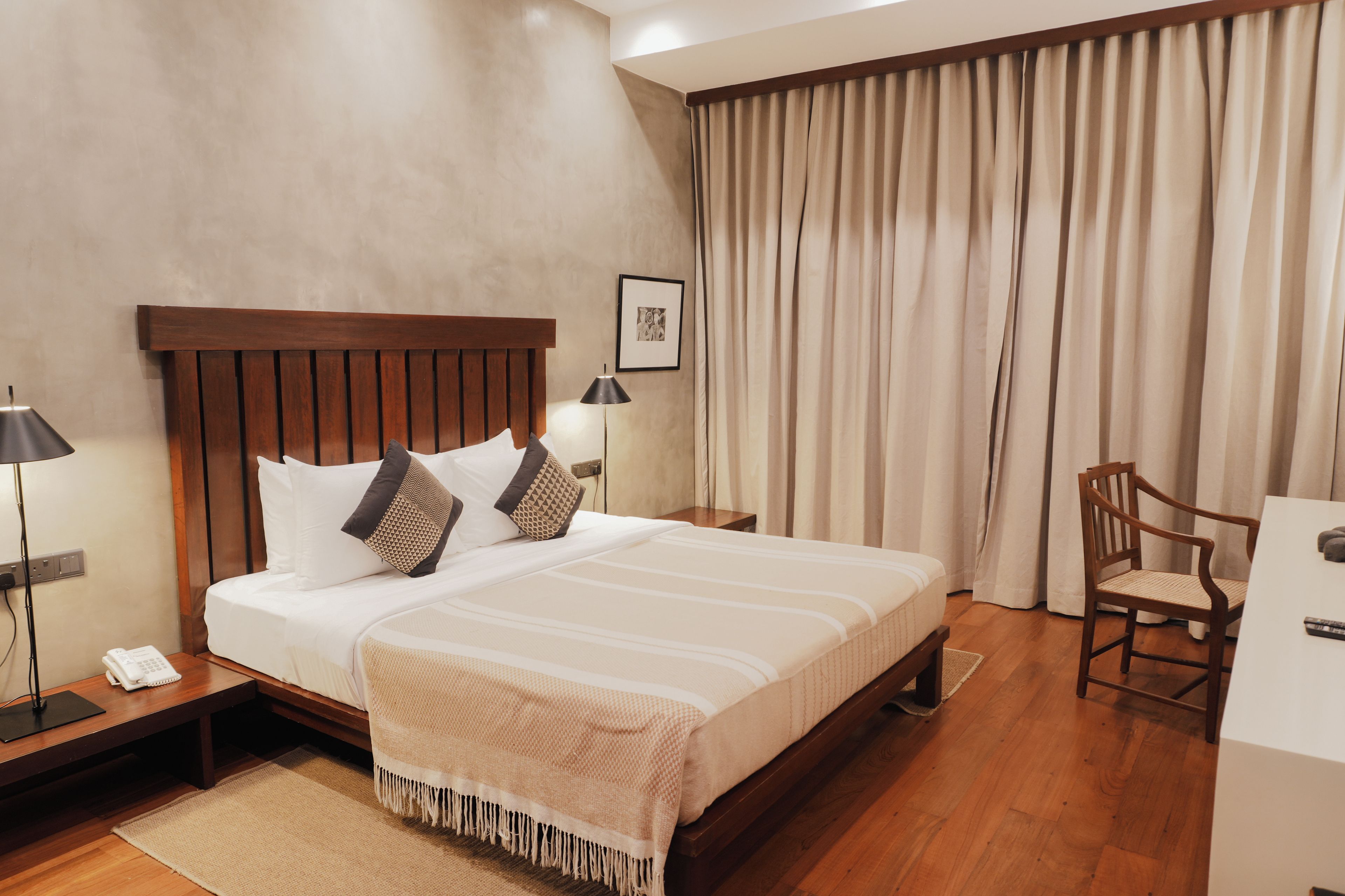 Habitación de hotel con cama de madera, escritorio, silla, lámpara y un amplio ventanal con cortinas.