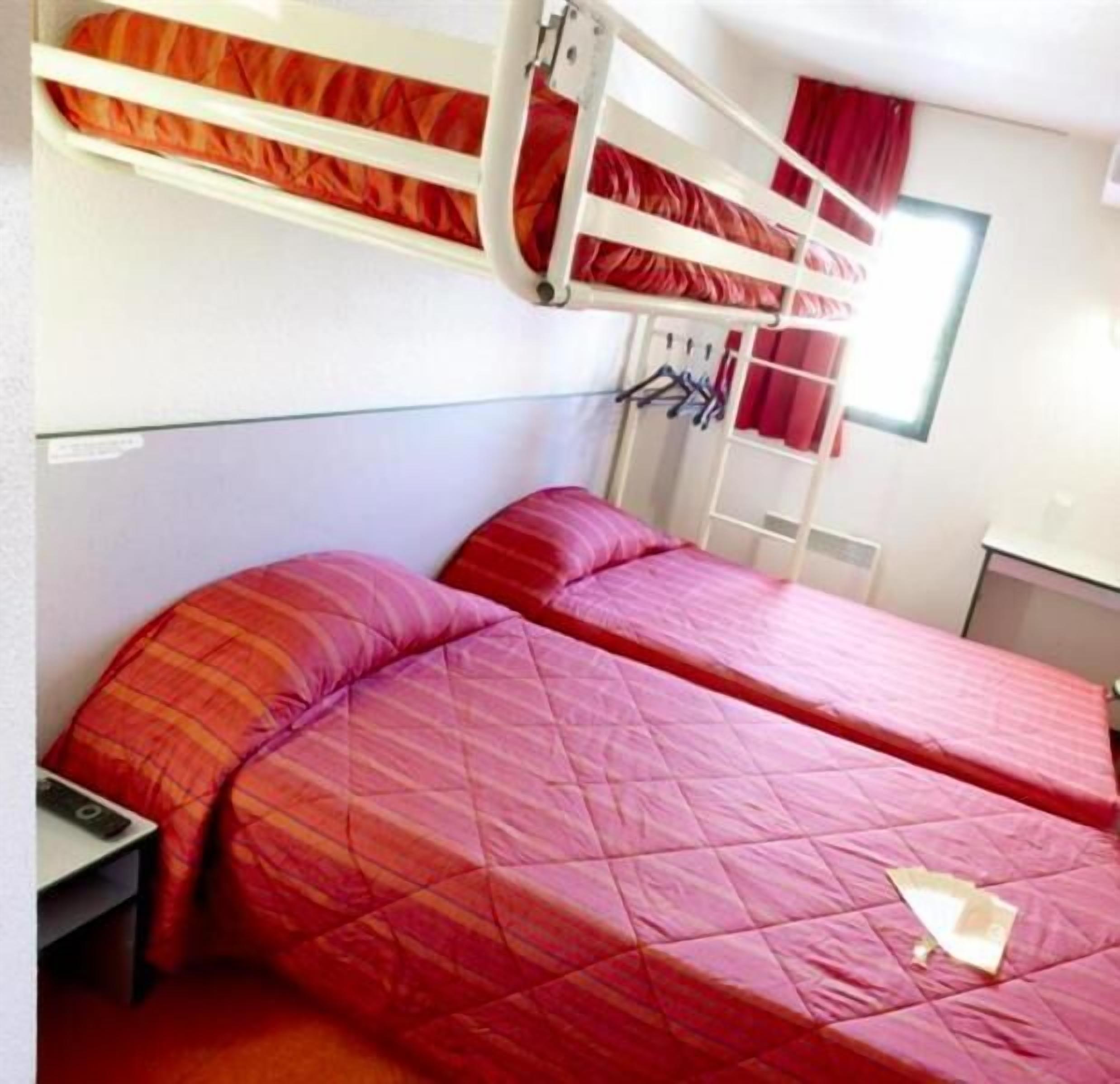 A bunk bed room with two beds, each with red bedding. There is a small table and a chair in the corner.