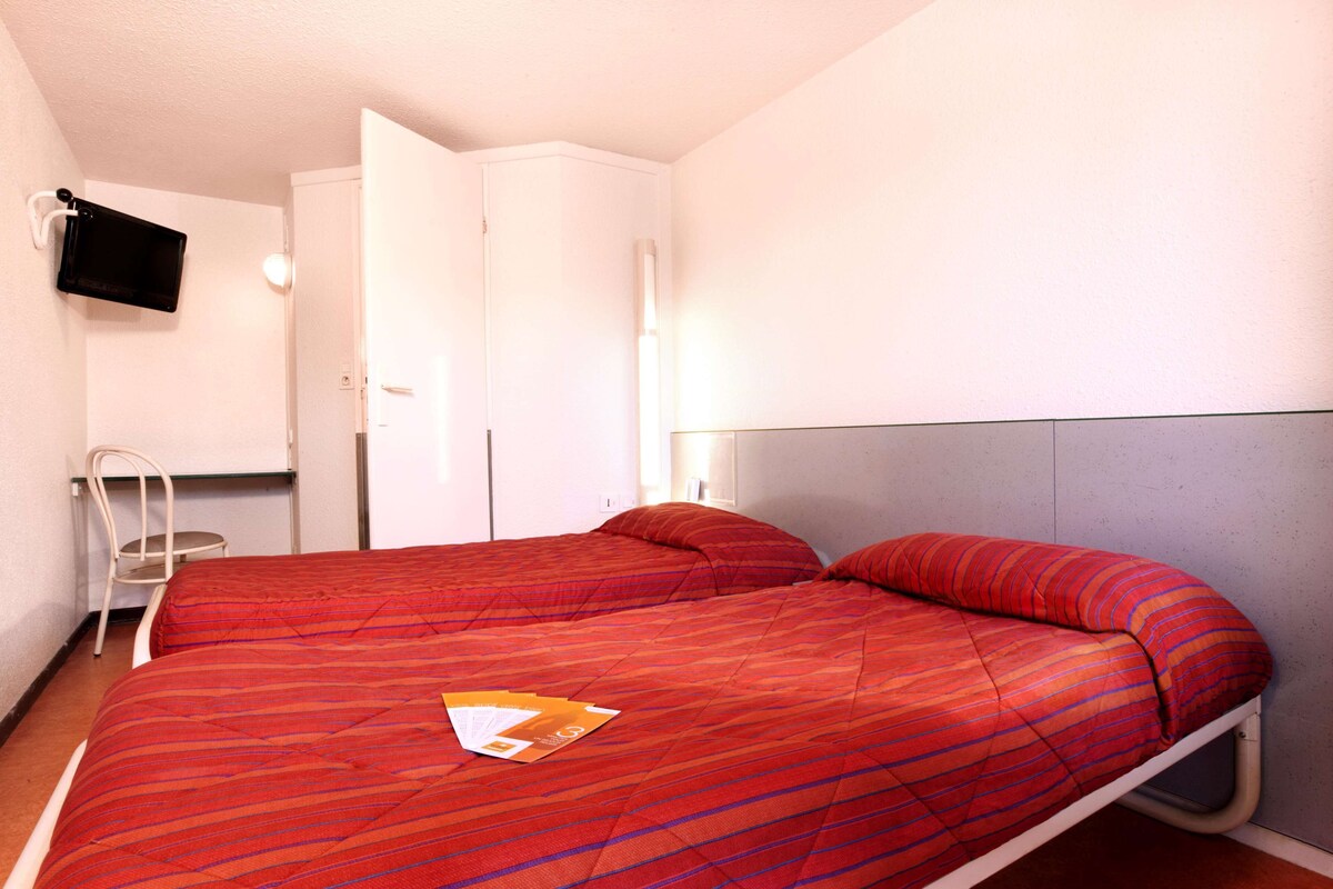 A single-bed room with a red-striped bedspread, a flat-screen TV mounted on the wall, a white wardrobe, and a chair.
