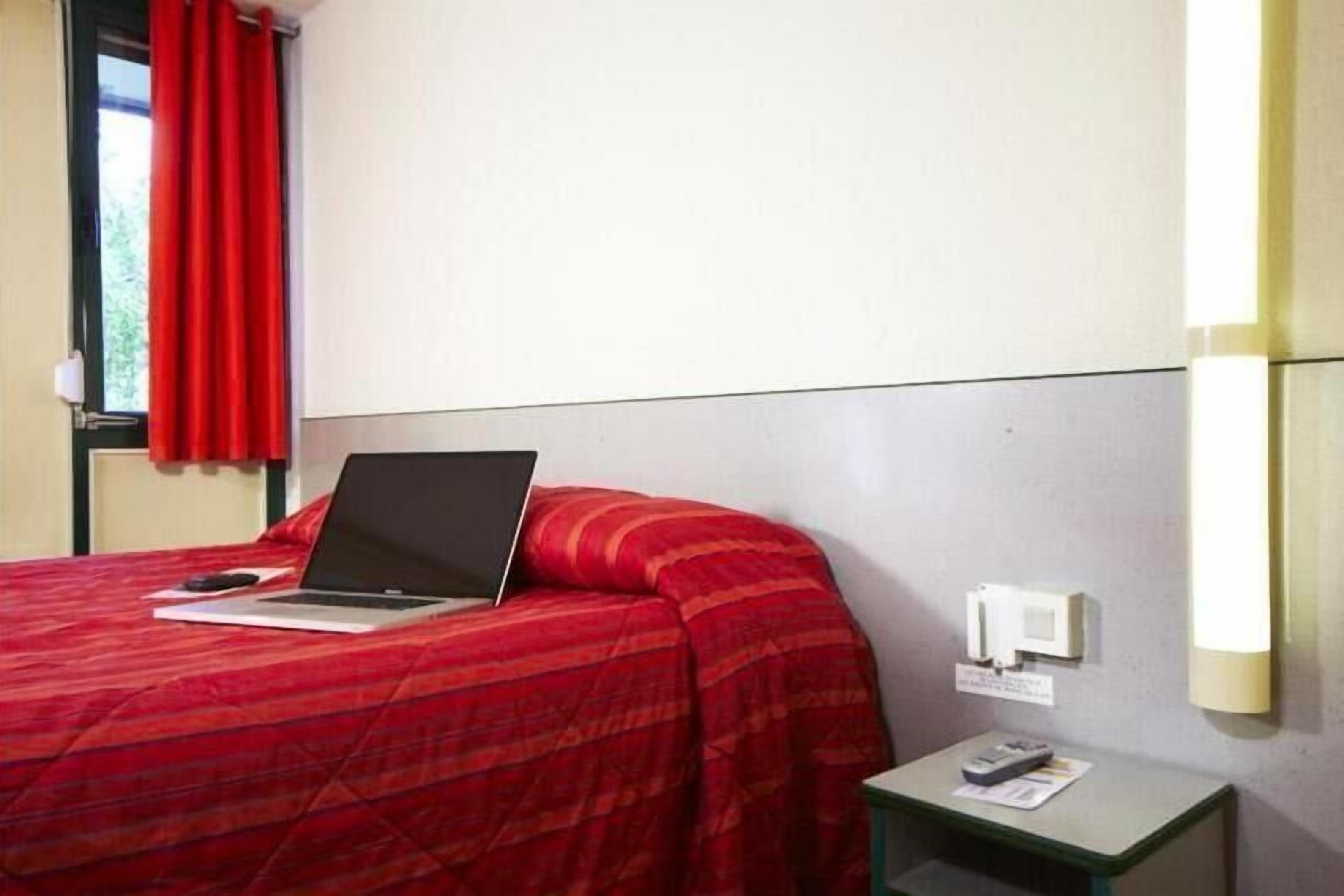 Hotelkamer met een bed, een laptop op het bed, een nachtkastje met een afstandsbediening en een wandlamp.