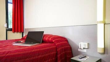 Hotelkamer met een bed, een laptop op het bed, een nachtkastje met een afstandsbediening en een wandlamp.