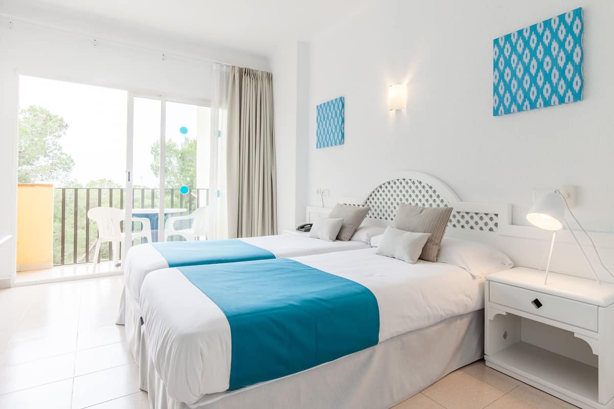 A hotel room with two beds, a balcony with chairs, and a view of greenery.