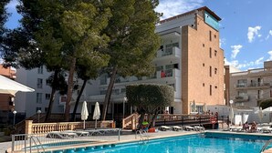 A swimming pool with lounge chairs and umbrellas, surrounded by trees and a multi-story building with balconies. - BLUESEA Costa Verde (Llucmajor)