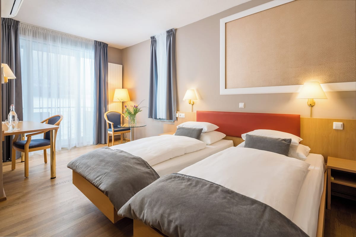 Ein Hotelzimmer mit zwei Betten, einem Schreibtisch, Stühlen und einem großen Fenster mit Vorhängen.