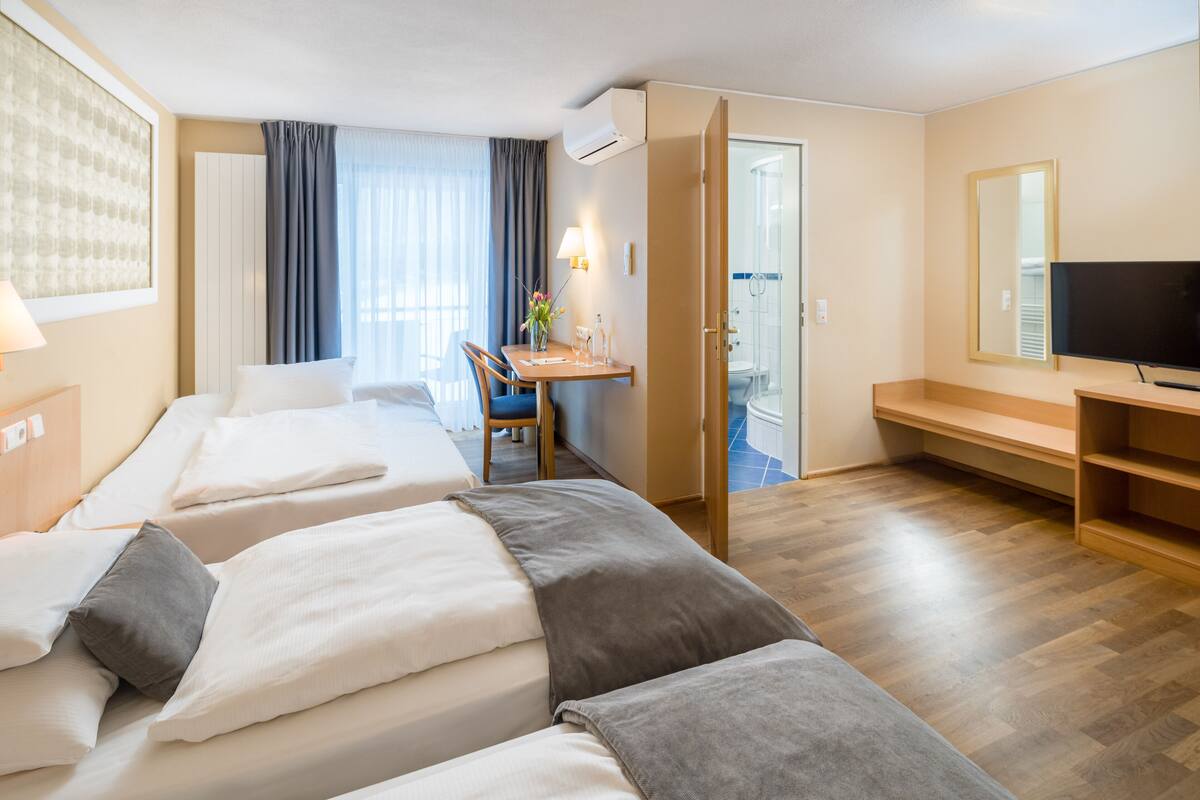 Ein Hotelzimmer mit zwei Betten, einem Schreibtisch, einem Stuhl, einem Fernseher und einem durchgegangenen Tür sichtbar erkennbaren Badezimmer.